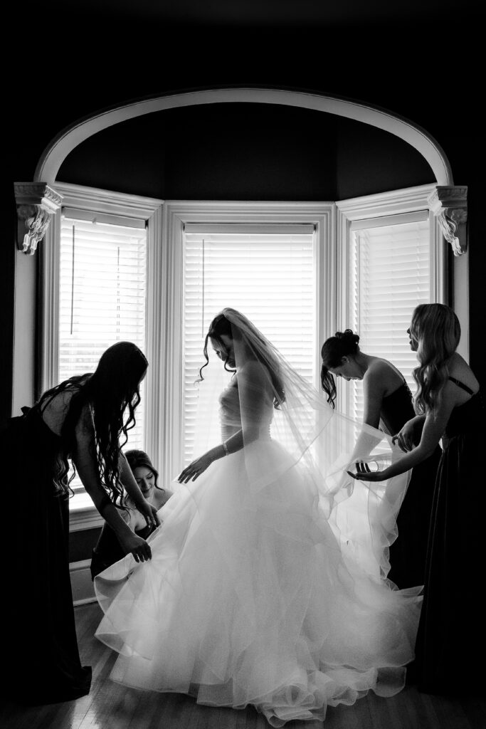 Bridesmaids fluffing ball gown in front of window